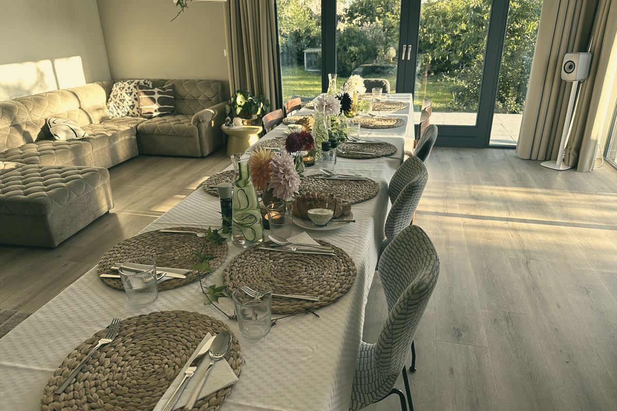 Lange gedeckte Tafel mit Blumen im Wohnraum, dahinter Glastüren zum Garten.