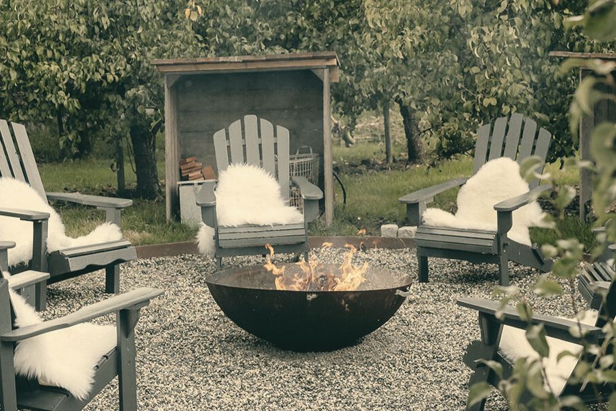 Brennende Feuerschale, ringsum Holzstühle mit Fellen unter Obstbäumen.