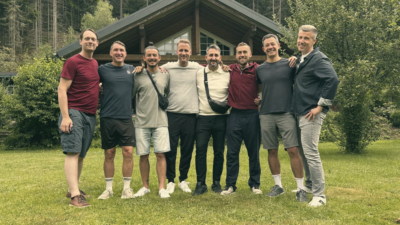 Acht Männer stehen Arm in Arm auf einer Wiese vor einer Holzhütte am Waldrand und lachen in die Kamera.